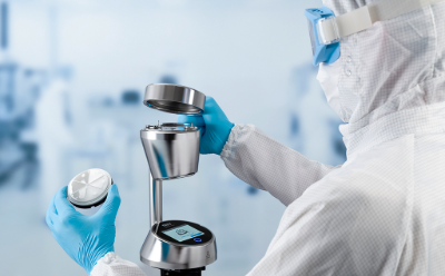A scientist in a protective suit and gloves examines a petri dish containing a sample, holding it up to the light against a blurred background of laboratory equipment.