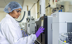 A female scientist wearing a white lab coat, a blue hairnet, safety goggles, and purple gloves is operating a sophisticated analytical instrument in a well-equipped laboratory. She is standing next to a black and silver machine with a touchscreen interface, carefully pressing the screen to adjust settings. The laboratory is filled with high-tech equipment, including a white gas chromatograph system with a series of sample vials neatly arranged in a circular tray. The background features an organized workspace with metallic and white surfaces, pipes, and analytical instruments mounted on the walls.