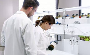 In a bright and modern laboratory, two scientists wearing white lab coats and safety goggles are engaged in an analytical procedure. The female scientist in the foreground, with dark brown hair tied back, is carefully holding a dark amber glass bottle with a black cap, tilting it slightly as she examines its contents. The male scientist standing beside her, with short brown hair and a beard, observes her actions attentively. The background features a well-organized laboratory bench with white shelves lined with multiple brown glass reagent bottles, some with blue and yellow caps. Stainless steel laboratory fittings, including a faucet and tubing system, are mounted on the wall. Large windows allow ample natural light to illuminate the workspace, emphasizing a clean, sterile environment.