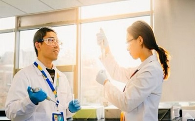Two scientists, a man and a woman, are working in a brightly lit laboratory with large windows allowing sunlight to stream in. The man, wearing safety goggles, a white lab coat, a blue lanyard with an ID badge, and blue nitrile gloves, is holding a pipette and attentively looking at his colleague. The woman, also in a white lab coat and safety glasses, is holding a test tube up to the light with a focused expression, carefully examining its contents. The background features laboratory benches with various equipment, and the golden sunlight casts a warm glow on the scene, emphasizing the collaborative nature of their work.