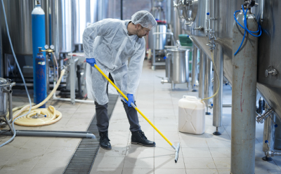 A worker in protective clothing is using a long-handled brush to clean the floor in a food processing facility. The background shows various equipment and tanks, emphasizing the importance of hygiene monitoring in food testing