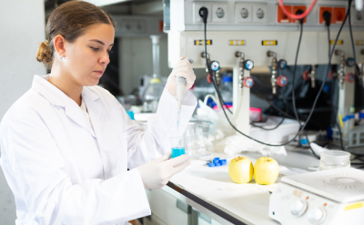 A laboratory technician in a white lab coat is preparing samples for food testing. The technician is holding a pipette and working at a lab bench.