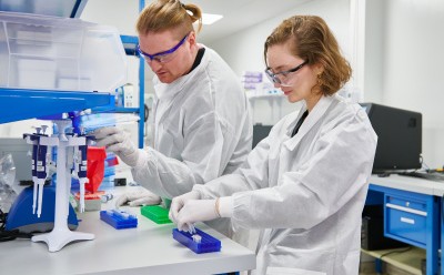 两名身穿白大褂的实验室技术人员正在实验台上工作，使用科学设备、制备样品。