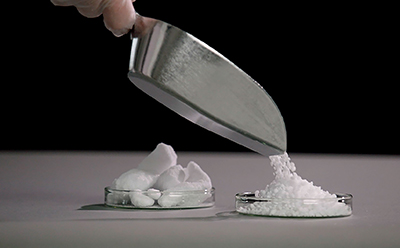 Stainless steel shovel pouring granulated raw material, demonstrating its reduced caking tendency and improved flow properties in comparison to bulk material