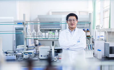 Scientist in pharma lab in a white lab coat stands with arms crossed in a laboratory setting, surrounded by various scientific equipment and glassware.