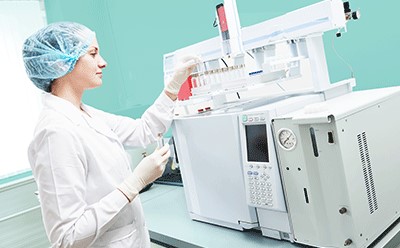 Female scientific researcher with lab coat, gloves and hair protection placing vial flask in gas chromatography instrument