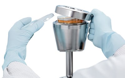 A close-up of a magnetic stainless steel lid with a handle being removed from a microbial air sampler, showing a 90 mm settle plate inside.