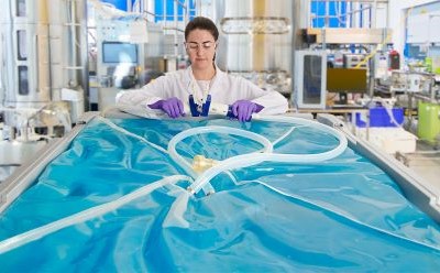 A person in a lab coat and purple gloves stands behind a large, transparent blue plastic container filled with liquid. Tubing is connected to the container, and the background shows various laboratory equipment and machinery.