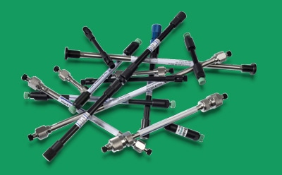各种 Supelco<sup>®</sup> 高效液相色谱柱和超高效液相色谱柱散落在绿色的表面上。这些色谱柱呈圆柱形，有金属表面，两端装有接头，代表了一系列满足不同分析需求的色谱柱技术。这些色谱柱大小不一，有些还贴有标明规格或型号的标签。
