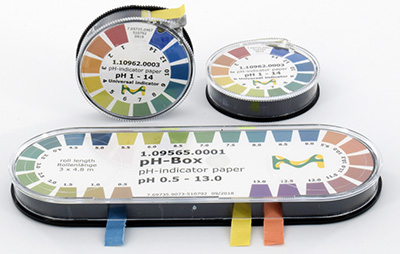 Three containers of pH indicator paper strips, each with a color-coded pH range chart on the lids. The containers are cylindrical, with one lying horizontally and two stacked vertically, all shown against a white background. Each container also has a small piece of tape in various colors to presumably mark different pH ranges.