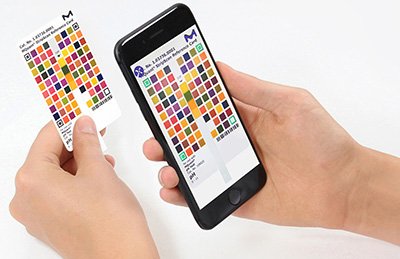 A person's hands holding a color chart card in one hand and a smartphone in the other. The smartphone screen displays a digital version of the same color chart, aligning the physical and digital formats for comparison or analysis. The background is white, emphasizing the items being held.