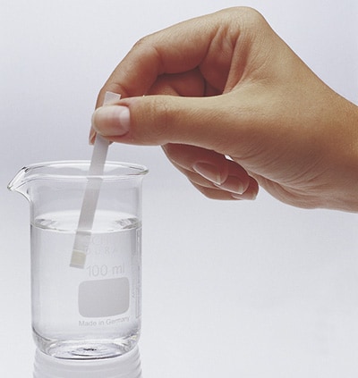 A hand dipping a test strip into a clear glass beaker filled with water. The beaker is marked with measurements indicating 100 ml and is labeled "Made in Germany." The action suggests a chemical or quality testing process.