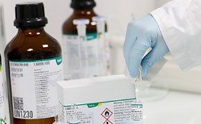 画面描绘了一个穿着白大褂、戴着手套的人在处理实验室柜台上的化学瓶和一个贴有危险标签的盒子，暗示了一个注重安全的环境。