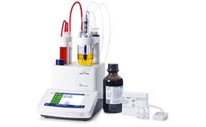 Modern laboratory titration setup featuring a digital titrator, various transparent reagent bottles with one containing a yellow liquid, and a dark brown bottle with a label, all connected by tubes, alongside a white tray with empty vials, indicating a precise chemical analysis process.