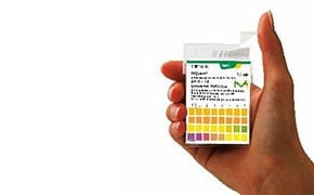 图片以白色为背景，描绘了一只手拿着 pH 试纸表，试纸表上有多个彩色方格，每个方格都标有从 0 到 14 的 pH 值。