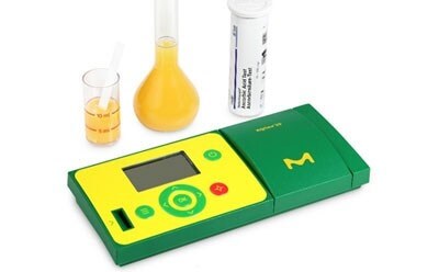 图片显示的是一个数字 pH 计，以绿色和黄色为主色调，配以一个盛有透明液体的烧杯、一个盛有黄色液体的烧瓶和一个空量筒，背景均为白色。