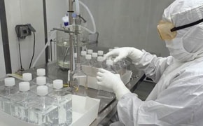 Technician in a cleanroom suit handling liquid cell culture media in a bioprocessing laboratory, with neatly arranged rows of sealed bottles containing the media and essential lab equipment in the background, indicating a sterile and controlled environment for bioprocessing applications.