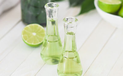 The image depicts two small clear glass bottles filled with green lime juice on a white wooden surface, accompanied by sliced limes and a bowl of whole limes in the background, suggesting freshly squeezed juice.