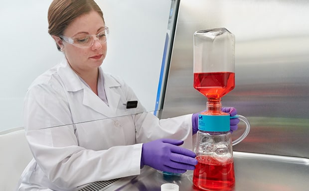 Sterile Filtration Devices A woman in a white lab coat, working with laboratory flask containing a red liquid, which is connected to another piece of equipment with blue liquid at the bottom.