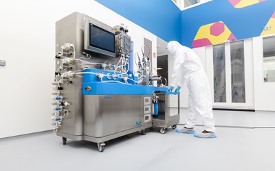 A technician in a white protective suit and blue shoe covers is operating a large, stainless steel pharmaceutical machine with blue accents, which features various tubes and a digital screen. The background shows a brightly colored, modern laboratory environment with geometric patterns on the walls.