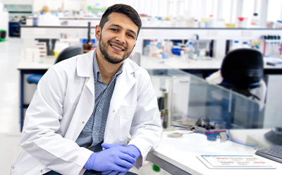 A scientist in a lab coat and gloves sits at a lab bench with various equipment in the background.