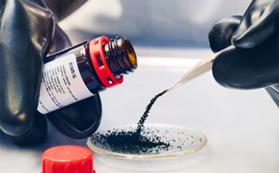 在实验室环境中，一名佩戴黑色手套的人员正用金属镊子将棕色玻璃瓶中的颗粒状深绿色或黑色粉末小心翼翼地转移至培养皿中。附近桌面上放置着一个红色瓶盖。