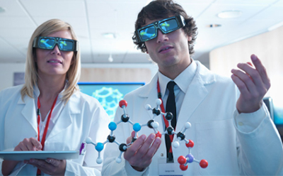 Two scientists, a woman and a man, stand side by side in a brightly lit, modern laboratory wearing white lab coats and large black 3D glasses. The woman, with straight blonde hair, holds a tablet in both hands and wears a red lanyard. The man has short brown curly hair and is holding a colorful molecular model made of plastic balls and sticks in black, blue, white, red, and purple, representing different atoms and bonds. He also wears a red lanyard and a name badge over a white shirt and black tie. Behind them, a digital screen displays a bright blue hexagonal molecular structure. The room is well-lit with soft white ceiling lights, pale wood walls, and a partially visible computer station in the background, giving the setting a clean and high-tech atmosphere focused on molecular research or visualization. 