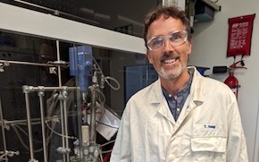 Haag stands in the lab in a lab coat and wearing safety glasses.