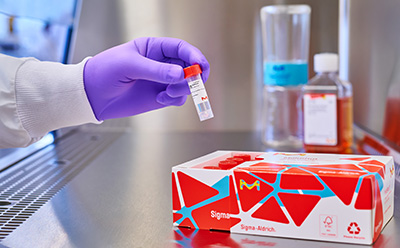 实验室场景：一只戴着手套的手握着一个红色瓶盖的小瓶，旁边放着一个几何图案的盒子，背景中还有其他实验室容器。