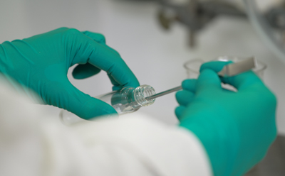 Two hands of an operator, clad in protective gloves, skillfully dispensing a fine white powder from a glass vial using a spatula.