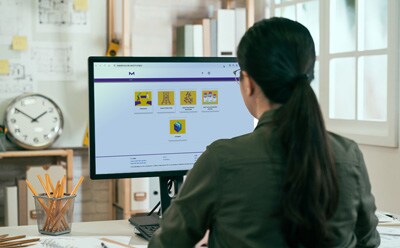 A woman with dark hair in a ponytail sits at a desk, looking at a computer monitor displaying a colorful dashboard with six yellow tiles.