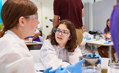 两个孩子穿着防护服，戴着手套，正在进行科学实践活动。一个孩子拿着一个容器，正在向另一个孩子演示什么，而另一个孩子则聚精会神地听着，背景中还能看到几个成年人和其他参与者，营造出一种互动和教育环境。