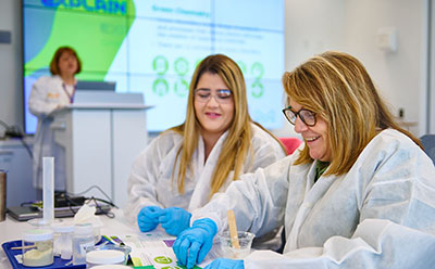Members of the Beyond Benign team are engaged in a scientific experiment at a table filled with various lab materials. In the background, a presentation screen displays information related to their work.