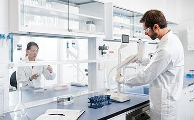 A laboratory scene showing two scientists in white lab coats working with lab equipment, including two Q-POD ultrapure water dispenser and various glassware.