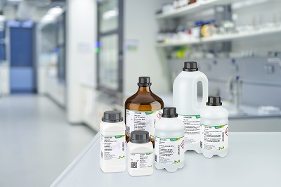 A variety of laboratory Emsure solvent bottles, labeled with product information, placed on a white counter in a modern laboratory setting. The bottles vary in size and shape, with both amber and white bottles visible, representing different chemical solvents typically used in analytical and research labs. The background features lab equipment and shelves