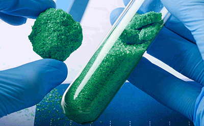 A gloved hand holding a blue powdery substance next to a test tube filled with similar blue granules, set against a laboratory background.