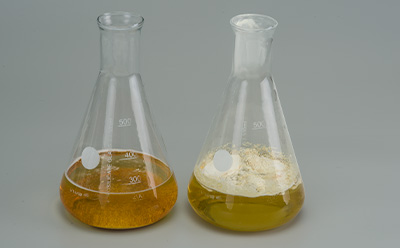 两只实验室烧瓶并排放置；左侧烧瓶盛有深琥珀色液体，右侧烧瓶则盛有浅黄褐色液体并带有些许沉淀，两者均置于灰色背景之上。
