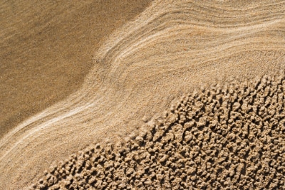 A close-up view of a sand, featuring textured patterns of wet and dry sand, with gentle waves creating smooth lines on the surface
