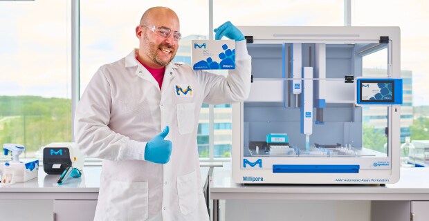 Scientist with SMC® Assay for AAW™ Workstation Scientist standing in front of AAW™ automated assay workstation holding up SMC® assay giving a thumbs up.