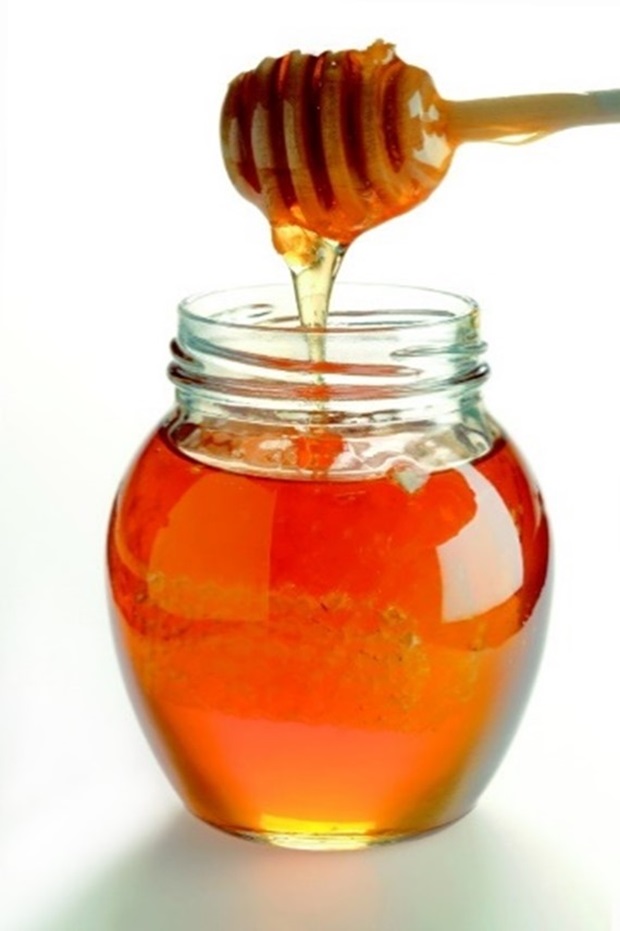 Glass jar filled with golden honey and honeycomb, with honey drizzling from a wooden dipper.
