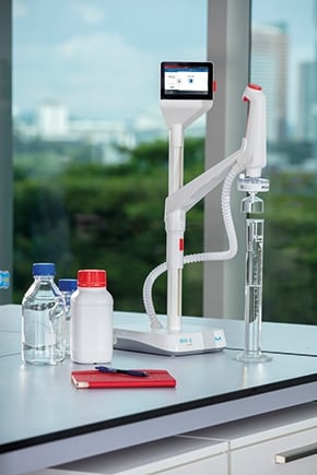 A laboratory setup featuring a digital dispensing device, glassware, and various containers on a countertop with a green outdoor view.