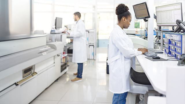 A laboratory scene showing a person in a lab coat working at a computer, while another individual stands nearby, both engaged in scientific tasks.