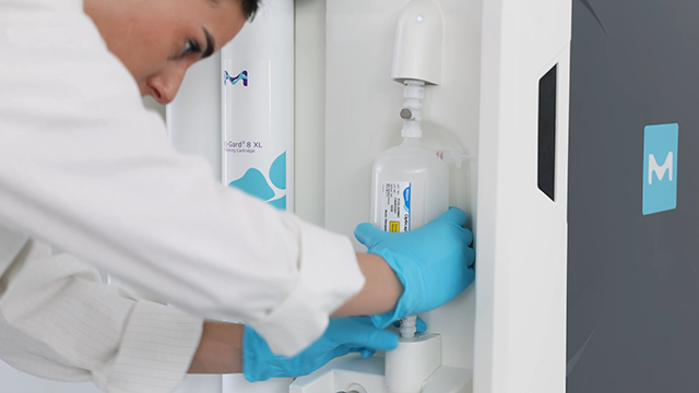 A person in a lab coat and blue gloves is carefully placing a bottle into a laboratory device, emphasizing precision in a clean environment.