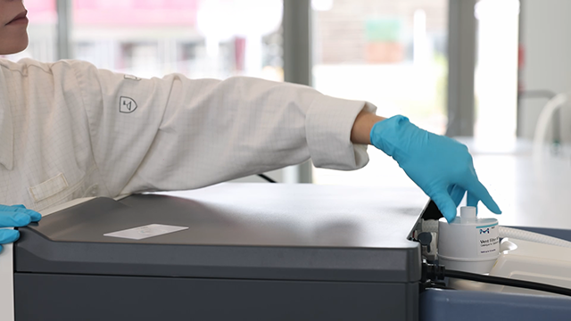 A person in a lab coat and blue gloves is placing a small container onto a laboratory instrument. Natural light illuminates the workspace.